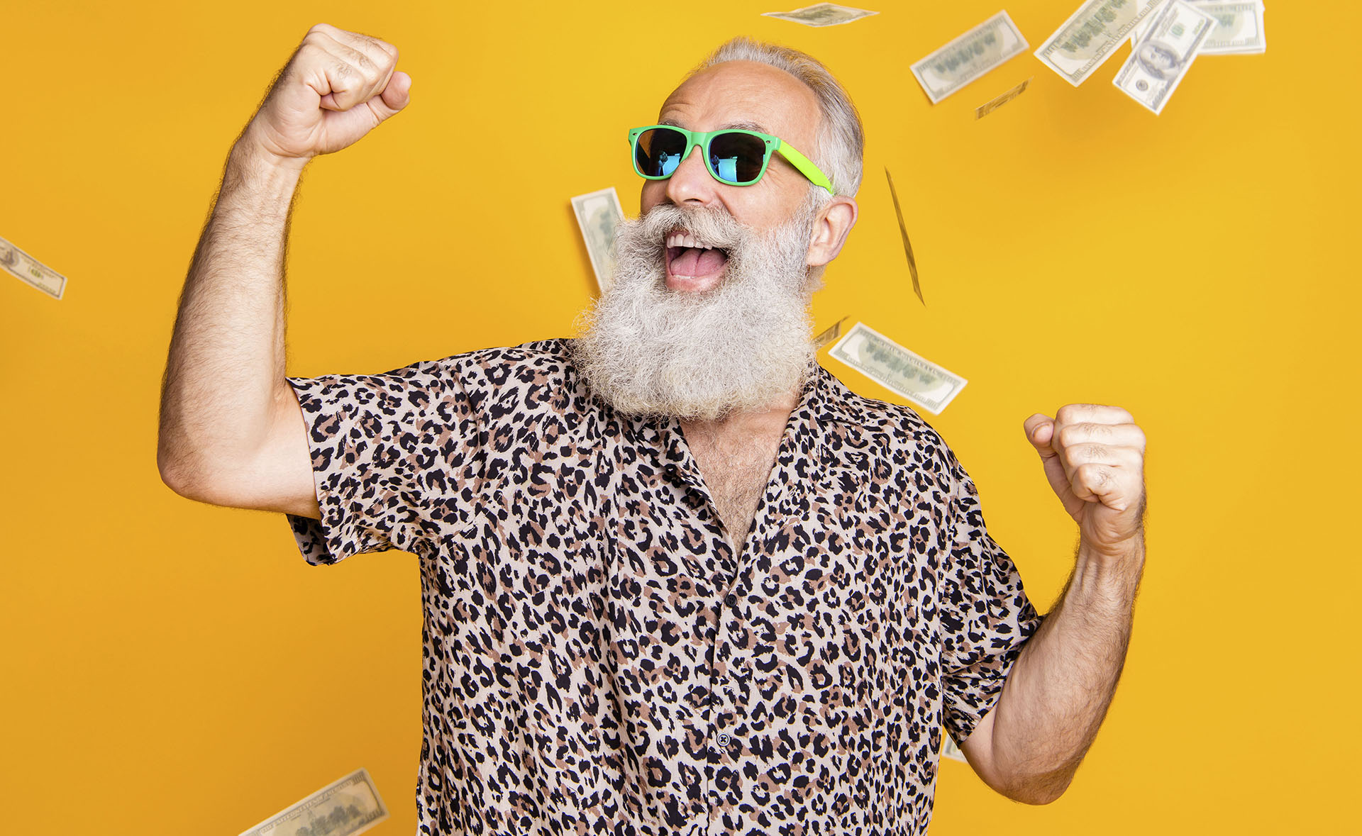 Portrait of crazy retired funky bearded old man with eyeglasses eyewear raise his fists scream yeah celebrate victory look at money falling wearing leopard shirt isolated over yellow background Älterer Hippster jubelt im Geldregen